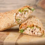 A warm, cheesy garlic chicken wrap filled with fresh vegetables, served on a wooden cutting board, with a side of dipping sauce.