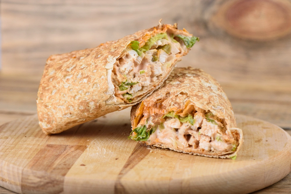 A warm, cheesy garlic chicken wrap filled with fresh vegetables, served on a wooden cutting board, with a side of dipping sauce.