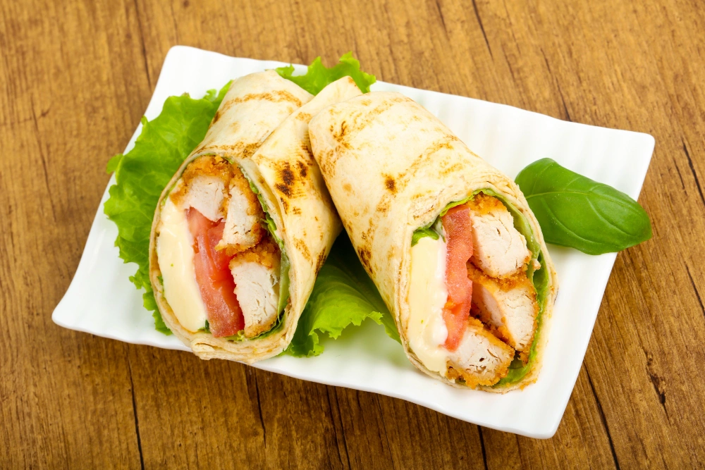 A warm, cheesy garlic chicken wrap filled with fresh vegetables, served on a wooden cutting board, with a side of dipping sauce.