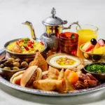 A colorful Arabic breakfast spread featuring a plate of Ful Medames made with fava beans, garlic, and lemon, served with warm pita bread. Accompanying the dish is creamy Labneh topped with olive oil and za'atar, alongside fresh vegetables and olives. Sweet Baklava drizzled with honey, fresh fruit, and a bowl of honey-sweetened porridge complete the meal, showcasing a delightful mix of flavors and textures.