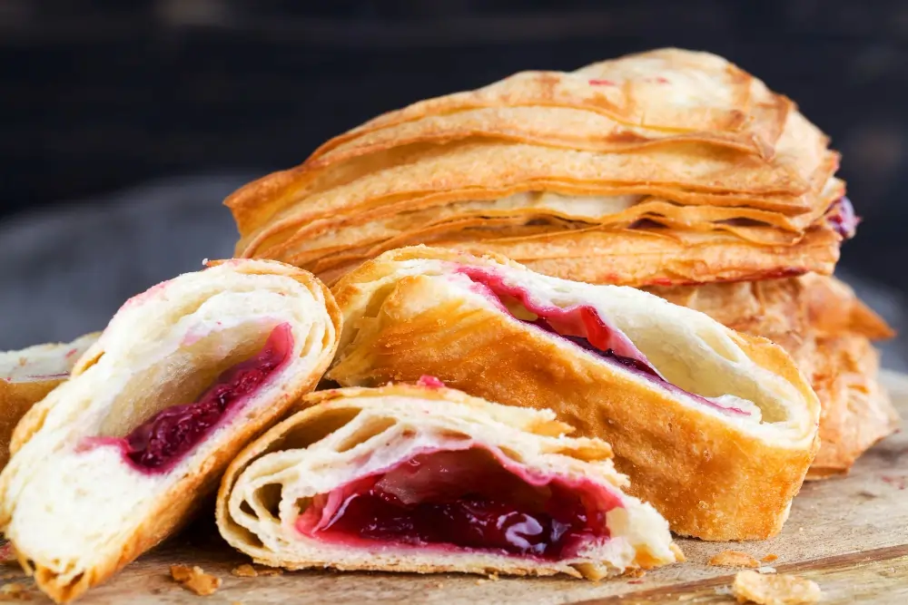 A freshly baked Breakfast Puff Pastry filled with sweet fruits and drizzled with icing, served on a wooden board alongside a cup of coffee and fresh berries, showcasing its flaky texture and inviting appearance.